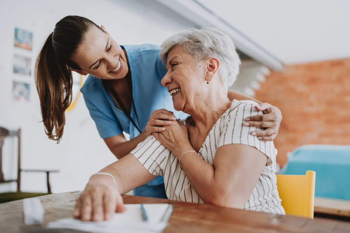 Caregiver smiling with her arm around a senior woman at a table